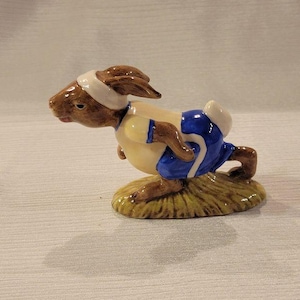 May include: A brown and white ceramic figurine of a rabbit wearing a blue and white running outfit. The rabbit is running on a green base.