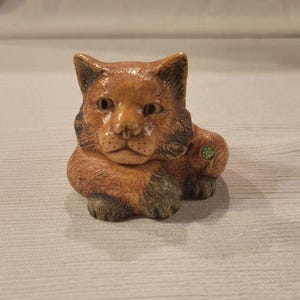 May include: A detailed, hand-carved figurine of a resting fox. The fox is primarily brown with darker accents around the ears and paws. A small green floral design is on its side. The figurine is resting on a light-colored surface.
