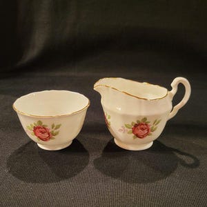 May include: A white porcelain creamer and sugar bowl set with a floral design featuring pink roses and green leaves. The set has a gold trim around the edges.