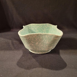 May include: A light green ceramic bowl with a speckled white pattern. The bowl has a unique, scalloped rim and a pedestal base. The bowl is likely vintage and suitable for serving food or as a decorative piece.