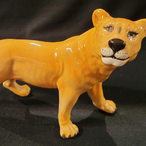 May include: A ceramic figurine of a lioness in a walking pose. The lioness is a glossy, golden-yellow color with a black nose and detailed facial features. The figurine is a decorative item.