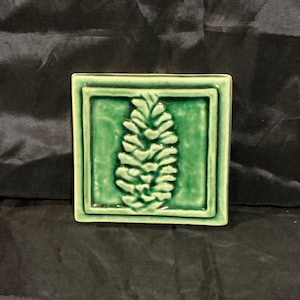 May include: A square ceramic tile featuring a glossy, deep green glaze. The center of the tile displays a raised, embossed relief of a pinecone. The tile has a raised border frame around the central square, creating a layered, dimensional appearance.