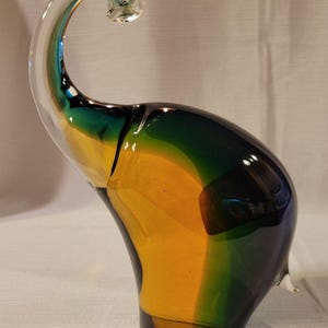 May include: A colorful glass elephant sculpture. The elephant is primarily yellow, green, and blue, with a clear, curled trunk. The sculpture has a smooth, glossy finish and is a decorative art piece.