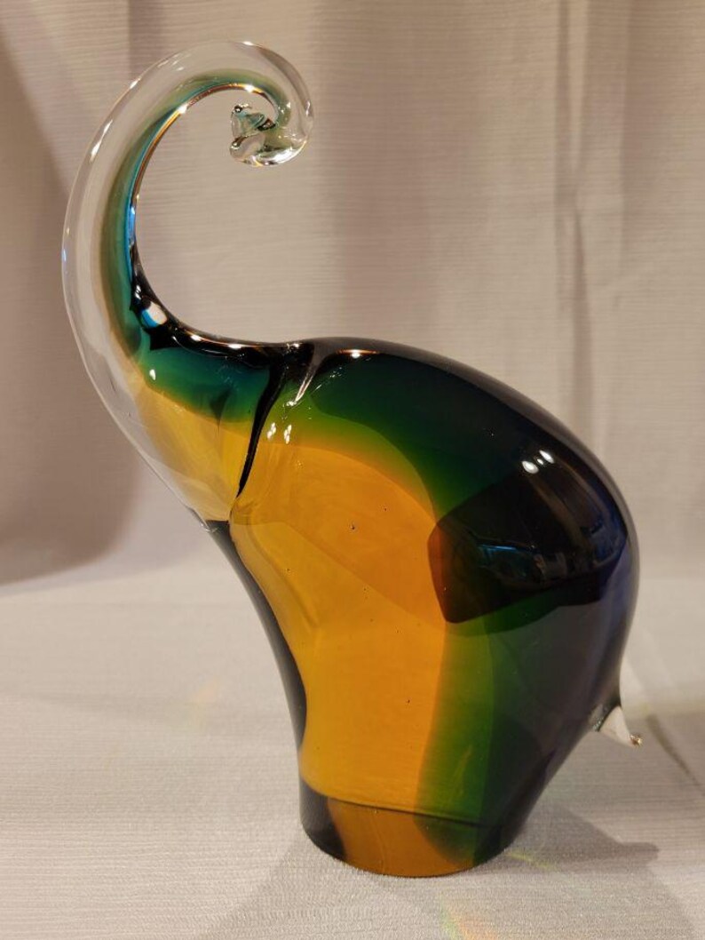 May include: A colorful glass elephant sculpture. The elephant is primarily yellow, green, and blue, with a clear, curled trunk. The sculpture has a smooth, glossy finish and is a decorative art piece.