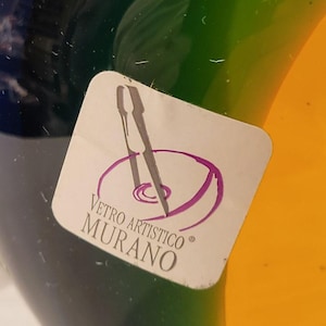 May include: Close-up of a colorful glass object with a sticker that reads "Vetro Artistico Murano." The glass has sections of blue, green, and yellow. The sticker features a purple design and text.