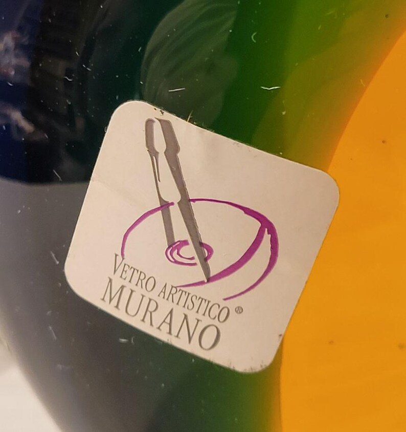 May include: Close-up of a colorful glass object with a sticker that reads "Vetro Artistico Murano." The glass has sections of blue, green, and yellow. The sticker features a purple design and text.