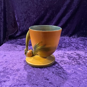 May include: A brown and green glazed ceramic vase with a pine cone handle. The vase has a wide base and a narrow opening.