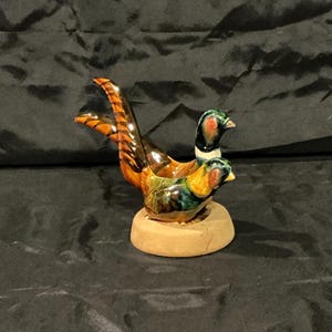 May include: A ceramic figurine of two pheasants, featuring detailed plumage in shades of brown, orange, green, and blue. The birds are perched on a small, round, light brown base. The figurine has a glossy finish.