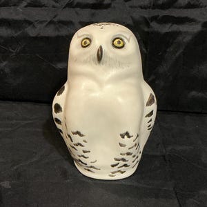 May include: A ceramic snowy owl figurine featuring a white body with dark brown speckled markings on the wings and chest. The owl has bright yellow eyes with black pupils and a dark brown beak, depicted in a forward-facing, upright posture.