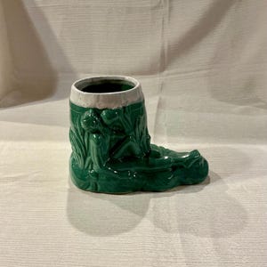 May include: Green ceramic boot-shaped mug with a white rim. The mug features a relief design of a reclining figure amidst foliage. The mug is approximately 6 inches long and 4 inches tall.