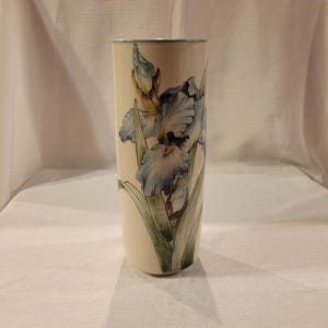 May include: A white ceramic vase with a blue and green floral design. The vase features a stylized iris flower with blue petals and green leaves.