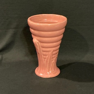 May include: A vintage, pink ceramic vase with a flared, ribbed body and a detailed base. The vase features stylized leaf-like accents on the side. The vase is a light, muted pink color.