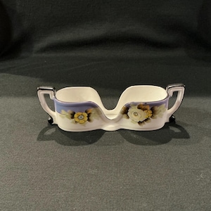 May include: A vintage ceramic double dish with a white base, adorned with floral designs in yellow and white. The dish features a lavender rim and black accents on the handles and feet. A decorative piece.