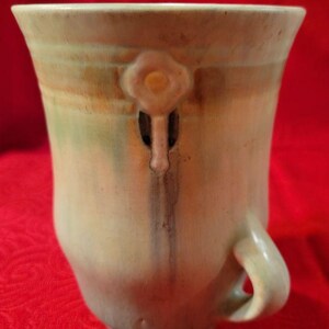 May include: A green ceramic vase with a light brown and pink floral design. The vase has a handle on one side and a unique design on the front.