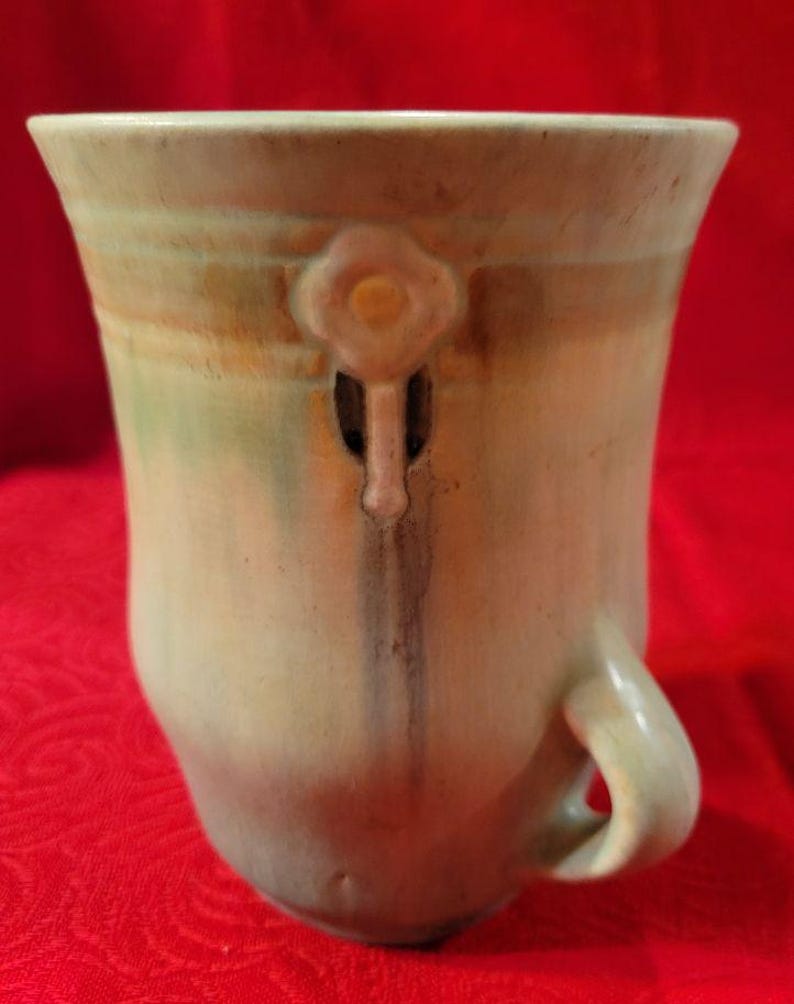 May include: A green ceramic vase with a light brown and pink floral design. The vase has a handle on one side and a unique design on the front.
