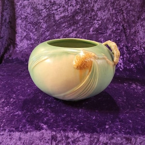 May include: A green and cream glazed ceramic vase with a pine cone design and two handles. The vase is on a purple velvet background.