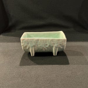 May include: A rectangular, light green ceramic planter with a floral design on the sides. The planter has three small, rectangular feet. The interior is a lighter shade of green. The planter is likely for small plants or succulents.