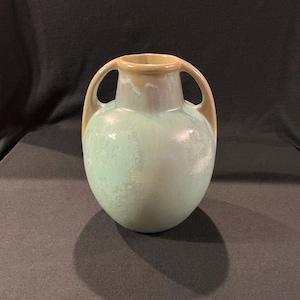 May include: A ceramic vase with a rounded body and two arched handles. The vase has a light green body with a tan rim and handles. The vase is likely used for decorative purposes.