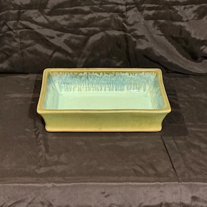 May include: A rectangular ceramic dish with a light green exterior and a smooth, pale blue interior. The upper rim features a decorative drip glaze effect where the blue glaze cascades down the inner walls of the vessel in vertical streaks.