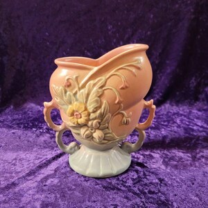 May include: A pink and cream ceramic vase with a floral design and two handles. The vase is shaped like a heart and has a pedestal base.