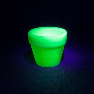 May include: A small, glowing green flower pot against a dark background. The pot has a classic shape with a slightly flared rim. The vibrant green color suggests it may be made of a material that glows under certain light conditions.
