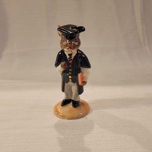 May include: A ceramic figurine of a rabbit wearing a black graduation cap and gown, holding a book. The rabbit has brown fur and a white shirt with a gold tie. The figurine is standing on a brown base.