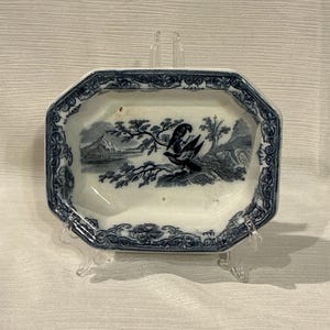 May include: A small, octagonal, antique dish with a blue and white bird and landscape design. The dish has a decorative border and is supported by a clear plastic stand. The bird is perched on a branch.
