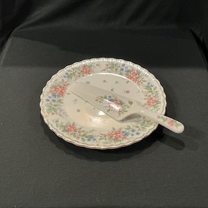 May include: A white ceramic cake plate and server set with a floral pattern in pink, blue, and green. The plate has a scalloped edge and a gold rim. The server is resting on the plate.