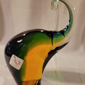 May include: A colorful glass elephant figurine with a curled trunk. The elephant is primarily yellow, green, and black, with a clear trunk. A sticker on the side reads "Vetro Artistico Murano".