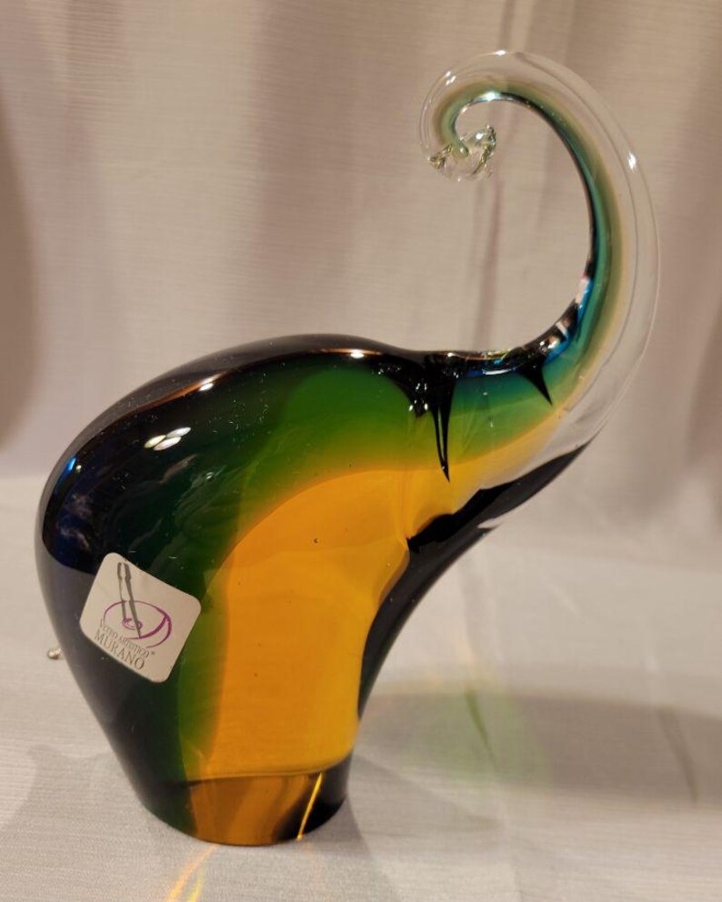 May include: A colorful glass elephant figurine with a curled trunk. The elephant is primarily yellow, green, and black, with a clear trunk. A sticker on the side reads "Vetro Artistico Murano".