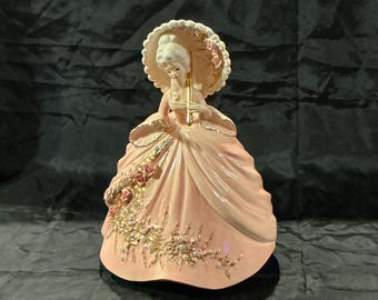 May include: A porcelain figurine of a woman in a pink dress, holding a parasol. The figurine has a detailed design with gold accents and floral embellishments. The woman's hair is styled in a classic updo.