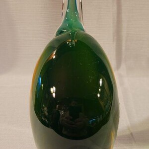 May include: A decorative glass bottle in a teardrop shape, primarily dark green with a clear, elongated neck. The bottle has a small, decorative element near the top and a pointed base. The glass reflects light, creating highlights.