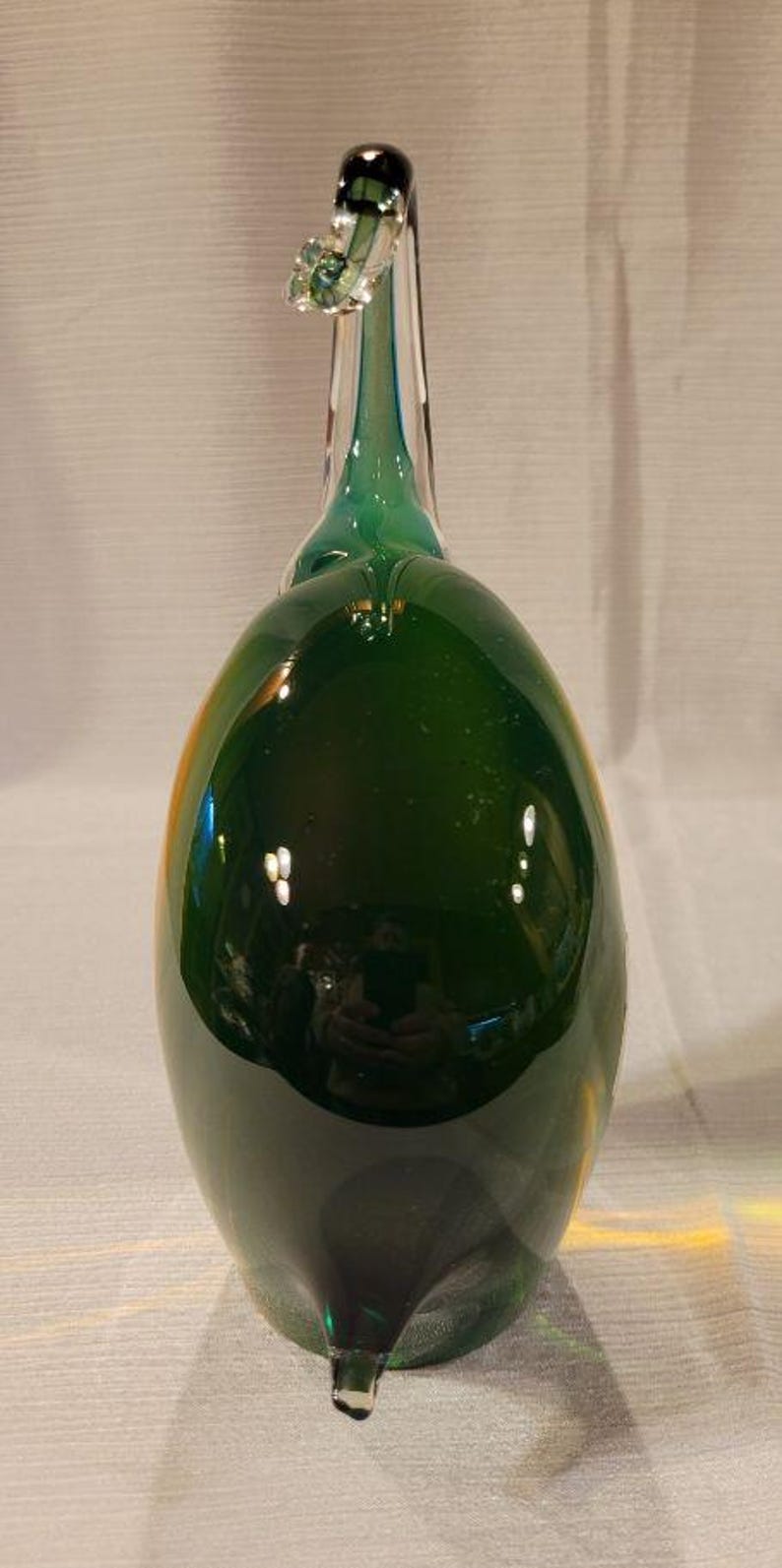 May include: A decorative glass bottle in a teardrop shape, primarily dark green with a clear, elongated neck. The bottle has a small, decorative element near the top and a pointed base. The glass reflects light, creating highlights.