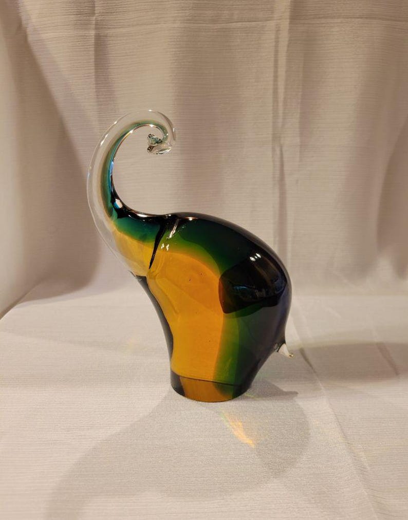May include: A colorful glass elephant sculpture with a curled trunk. The sculpture features a gradient of colors, including yellow, green, and blue. The elephant is a decorative art piece.
