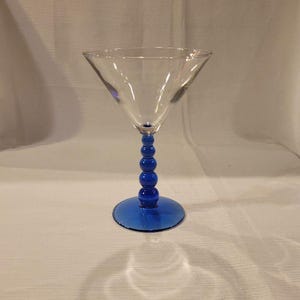 May include: A clear glass martini glass with a blue stem and base. The stem is made of stacked blue spheres. The glass has a wide, triangular bowl. The base is a flat, circular design. The glass is set against a white background.