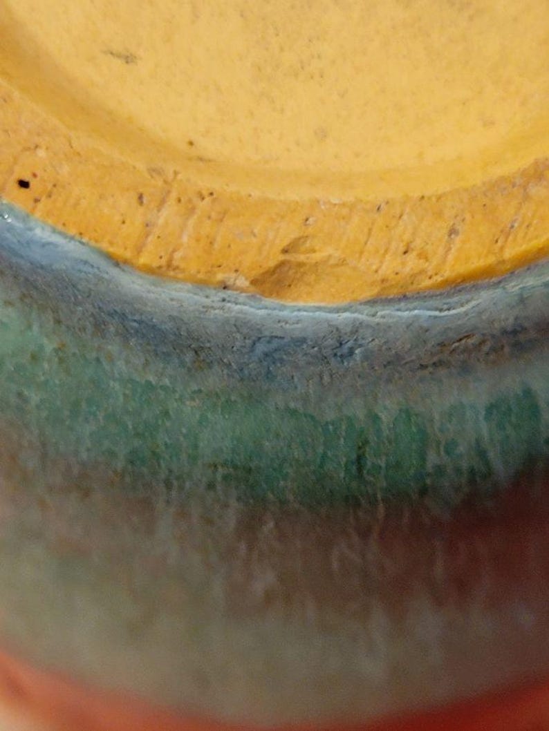 May include: Close-up of the bottom of a ceramic vase with a multi-colored glaze. The glaze is a gradient of orange, green, blue, and yellow.