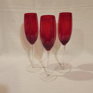 May include: Three red champagne flutes with clear stems and bases. The glasses are made of glass and are arranged in a row, with the light reflecting off the red color.