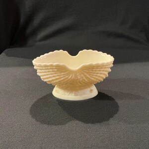 May include: Cream-colored, seashell-shaped dish with a scalloped rim and a pedestal base. The dish has a textured, ridged design, and the base has a decorative pattern. This decorative dish is suitable for serving small items.