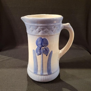 May include: A white ceramic pitcher with a blue and white floral design. The pitcher has a handle and a wide mouth.