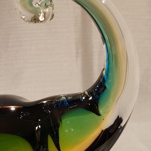 May include: A handcrafted glass sculpture with a curved, horn-like shape. The clear glass transitions to shades of green, yellow, and blue, with a black base. The sculpture has a unique, artistic design.