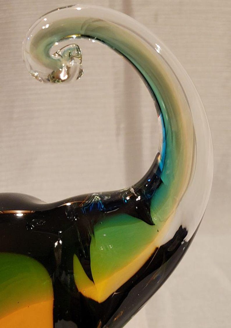 May include: A handcrafted glass sculpture with a curved, horn-like shape. The clear glass transitions to shades of green, yellow, and blue, with a black base. The sculpture has a unique, artistic design.