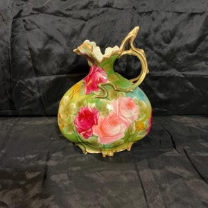 May include: A porcelain pitcher featuring a hand-painted floral design with vibrant red and soft pink roses against a green background. The vessel has a scalloped rim and a curved, gold-toned handle with ornate, flowing details.