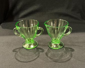 May include: Two vintage green glass cream and sugar set. The sugar bowl has two handles, and the creamer has a spout. Both pieces have a pedestal base and a ribbed design. The glass has a bright, neon green color.