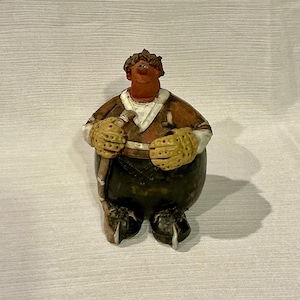 May include: A whimsical figurine of a person in a brown and white hockey uniform, holding a hockey stick. The figure has yellow gloves and black boots. The figurine has a smiling face and a round body.