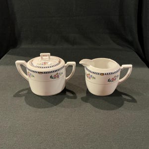 May include: A white ceramic sugar bowl with a lid and a creamer. Both pieces have a floral design with blue and black trim. The sugar bowl has two handles and a small handle on the lid. The creamer has a handle and a spout.