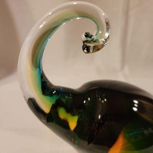 May include: A handcrafted glass sculpture with a swirling, curved design. The sculpture features a clear, transparent top with streaks of blue, green, and yellow. The base is a dark, glossy green with hints of orange.