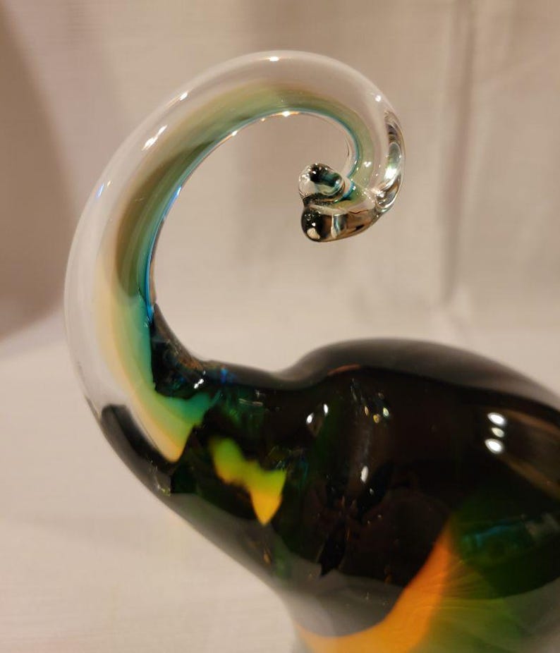 May include: A handcrafted glass sculpture with a swirling, curved design. The sculpture features a clear, transparent top with streaks of blue, green, and yellow. The base is a dark, glossy green with hints of orange.