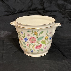 May include: A cream-colored, ceramic planter with a floral design. The planter features pink, blue, and yellow flowers with green leaves. It has gold trim around the top and base, and small handles on either side.