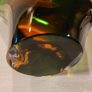 May include: A close-up of a glass vase with a wide base and a flared top. The glass is a dark, translucent color with streaks of orange, green, and yellow. The base is a solid, dark color, and the light reflects off the glass.