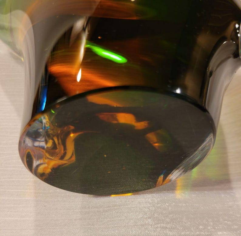 May include: A close-up of a glass vase with a wide base and a flared top. The glass is a dark, translucent color with streaks of orange, green, and yellow. The base is a solid, dark color, and the light reflects off the glass.